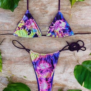 May include: A two-piece bikini set with a vibrant tie-dye pattern in shades of purple, pink, blue, and green. The triangle top and thong-style bottom feature thin, adjustable straps. The fabric has a shimmering, metallic finish.