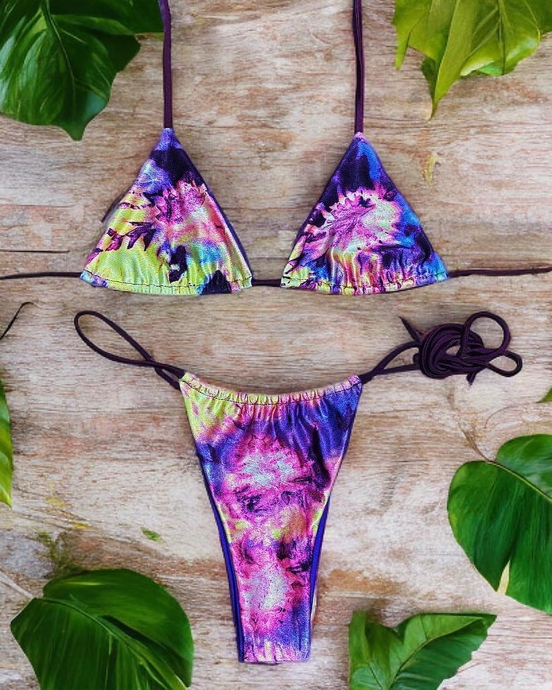 May include: A two-piece bikini set with a vibrant tie-dye pattern in shades of purple, pink, blue, and green. The triangle top and thong-style bottom feature thin, adjustable straps. The fabric has a shimmering, metallic finish.