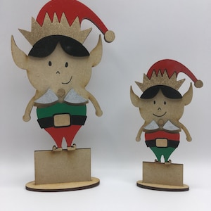Puede incluir: Dos adornos de elfo de madera, uno más grande que el otro. Ambos elfos llevan gorros rojos con coronas doradas, camisas verdes, pantalones rojos y zapatos negros. Los elfos están de pie sobre bases de madera.