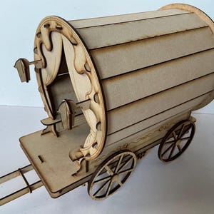 Puede incluir: Un vagón de madera en miniatura con una parte superior redondeada y ruedas de radios. El vagón presenta recortes decorativos y un diseño detallado, con una barra de tracción que se extiende desde la parte delantera. El vagón es de madera marrón claro.