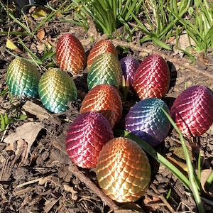 Puede incluir: Una colección de 11 huevos de dragón iridiscentes, cada uno con un degradado de color diferente, están esparcidos sobre una cama de mantillo marrón y hierba verde. Los huevos están hechos de un material brillante y texturizado que se asemeja a escamas de dragón.
