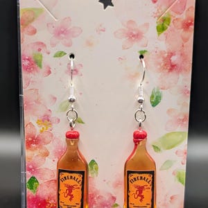 May include: A pair of dangle earrings featuring miniature Fireball whiskey bottles. The earrings have silver-colored hooks and red bottle caps. The bottles are orange with black and yellow labels. The earrings are displayed on a floral card.