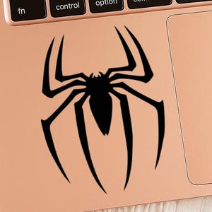 Pu&ograve; includere: Un adesivo nero a forma di ragno &egrave; applicato a un laptop color oro rosa. Il design del ragno &egrave; un'illustrazione stilizzata con otto zampe e un corpo centrale. La tastiera e il touchpad del laptop sono visibili.