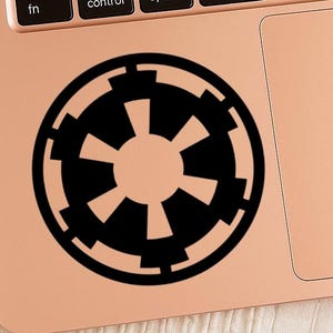 May include: A black vinyl sticker featuring the Galactic Empire symbol, a circular design with a central starburst pattern. The sticker is placed on a rose gold laptop, with the keyboard visible in the background.