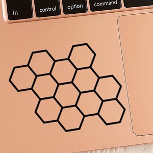 May include: A black honeycomb pattern sticker on a rose gold laptop. The sticker is composed of interconnected hexagonal shapes, creating a geometric design. The laptop keyboard and trackpad are visible.