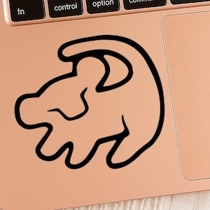 May include: A black outline sticker of a lion cub's head, placed on a rose gold laptop. The sticker's design is a simplified, cartoon-like illustration of the cub's profile. The laptop's keyboard and trackpad are also visible.