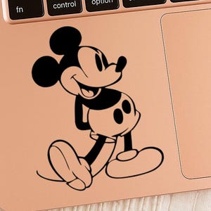 May include: Black and white Mickey Mouse decal on a rose gold laptop. The cartoon character is smiling and walking, with his iconic large ears and white gloves. The laptop keyboard and trackpad are visible.