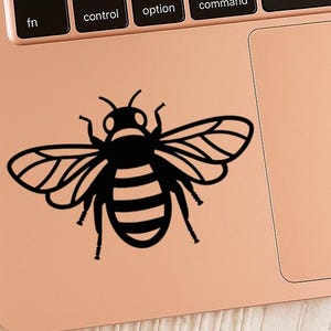 May include: A black and white bee sticker on a rose gold laptop. The bee is a stylized illustration with detailed wings and a striped abdomen. The laptop keyboard and trackpad are visible.