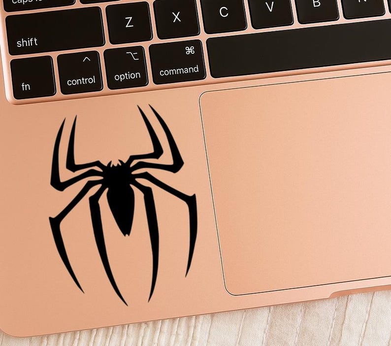 Pu&ograve; includere: Un adesivo nero a forma di ragno &egrave; applicato a un laptop color oro rosa. Il design del ragno &egrave; un'illustrazione stilizzata con otto zampe e un corpo centrale. La tastiera e il touchpad del laptop sono visibili.