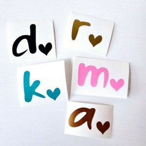 May include: Five white square stickers with a letter and a heart. The letters are black, gold, teal, pink, and brown. Each letter is paired with a heart in a matching color.