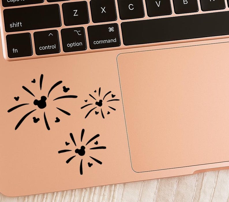 K&ouml;nnte beinhalten: Schwarze, gestanzte Vinylaufkleber mit einem stilisierten Feuerwerksdesign mit Mickey Mouse-K&ouml;pfen und Herzen. Die Aufkleber sind in einer Dreiergruppe auf einer ros&eacute;goldenen Laptop-Oberfl&auml;che angeordnet.