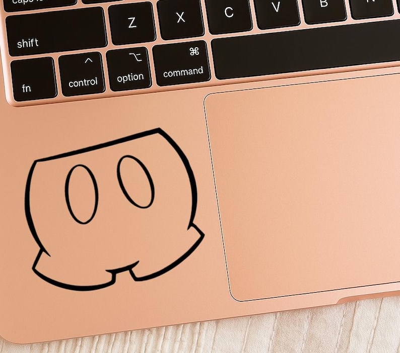 Puede incluir: Una pegatina en blanco y negro de pantalones cortos de dibujos animados con dos formas ovaladas. La pegatina est&aacute; colocada en un port&aacute;til de color oro rosa, cerca del teclado y el panel t&aacute;ctil.