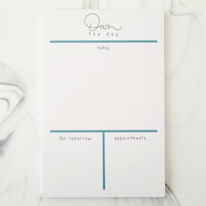 May include: White notepad with a blue line across the top and a blue line and vertical line across the bottom. The text "Own the day" is written in black cursive at the top. The text "today" is written in black below the top line. The text "for tomorrow" is written in black to the left of the vertical line. The text "appointments" is written in black to the right of the vertical line.