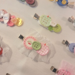 Pode incluir: Uma coleção de presilhas de cabelo com vários elementos decorativos, incluindo botões coloridos em tons de rosa, verde, azul e amarelo, pequenos laços brancos e detalhes em tule branco transparente fixados em presilhas de metal tipo jacaré.
