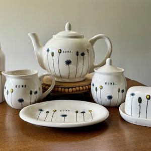 May include: A ceramic tea set in cream with a hand-painted floral pattern of thin stems and blue and yellow flowers. Each piece features the word HOME. in black text. The set includes a teapot, a lidded canister, a creamer, a lidded sugar bowl, a plate, and a butter dish.