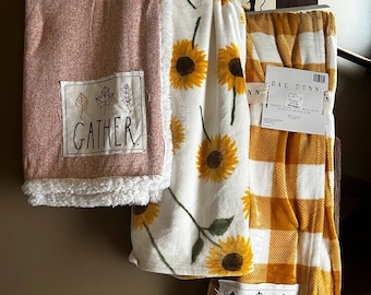 May include: Three throw blankets with a fall theme. The blankets are made of soft, plush fabric and feature a variety of fall-inspired designs, including sunflowers, plaid, and a gather design with leaves.