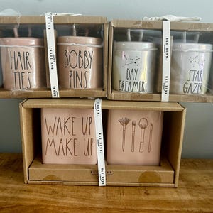 Könnte beinhalten: Eine Sammlung von Rae Dunn Keramikbehältern in Kartons. Die Behälter sind in Rosa- und Weißtönen gehalten und tragen schwarzen Text mit der Aufschrift "HAIR TIES", "BOBBY PINS", "DAY DREAMER", "STAR GAZER", "WAKE UP MAKE UP" und Make-up-Pinsel-Illustrationen.