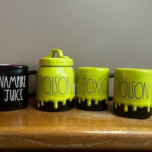 May include: Four Halloween-themed ceramic mugs and a jar. The black mug reads "VAMPIRE JUICE" in white. The jar and two mugs are bright green with black dripping paint and the words "POISON" and "TOXIC". The mugs have black handles.