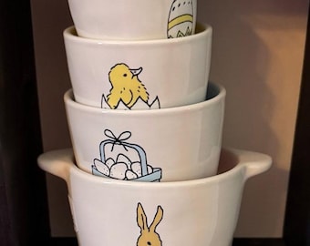 May include: A stack of four white ceramic bowls with Easter-themed illustrations. The bowls feature a yellow chick, a bunny, an Easter egg, and a basket of eggs. Each bowl has a different design, perfect for serving snacks or small portions.