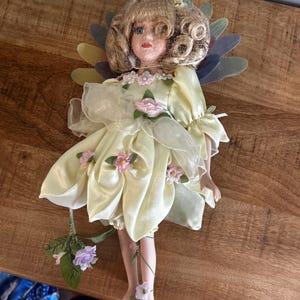 Pode incluir: Uma boneca de porcelana com cabelo loiro encaracolado veste um vestido de cetim amarelo pálido adornado com pequenas flores de tecido rosa. A boneca apresenta asas translúcidas multicoloridas, um adorno floral e segura um caule com flores roxas.