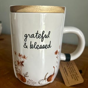 Może przedstawiać: Biały ceramiczny kubek z drewnianą pokrywką, ozdobiony czarnym napisem grateful & blessed. Kubek posiada pomarańczowe i brązowe ilustracje dyń, liści oraz polnych kwiatów. Do uchwytu przymocowana jest brązowa papierowa etykieta.