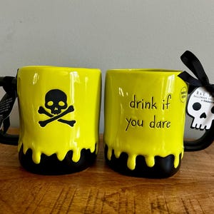 Puede incluir: Dos tazas de cerámica de color amarillo brillante con asas negras y un diseño de goteo negro en la base. Una taza presenta un gráfico de calavera y tibias cruzadas, mientras que la otra tiene el texto "drink if you dare" y una etiqueta de calavera.