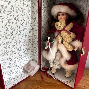 May include: A porcelain doll with brown hair wearing a red velvet coat with white faux fur trim and a matching hat. The doll holds a small tan teddy bear and a miniature wreath. It is displayed inside a red box lined with white floral paper.
