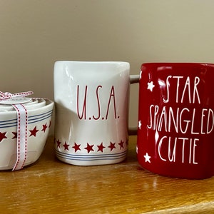 May include: A set of white ceramic measuring bowls with red stars and blue stripes, tied with a ribbon. A white mug with "U.S.A." in red lettering and a red mug with "STAR SPANGLED CUTIE" in white lettering and star accents.