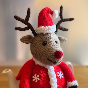May include: A knitted reindeer tea cozy. The cozy is brown with a red nose, antlers, and a Santa hat. It is wearing a red coat with white snowflake accents and white trim. The cozy is designed to fit over a white teapot.