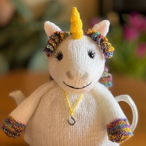 May include: White knitted teapot cozy shaped like a unicorn with a yellow horn, rainbow yarn mane, and a silver horseshoe charm.