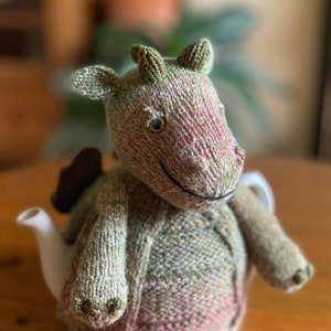 Peut inclure: Un chauffe-théière en forme de dragon tricoté en vert et rose avec un bec blanc et un visage souriant.
