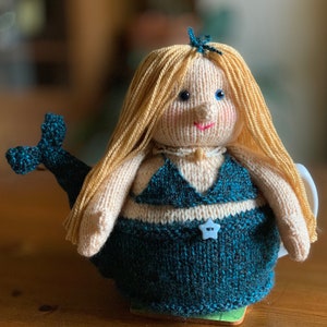 May include: A knitted mermaid teapot cozy with a teal tail, a white bikini top, and a blue star button. The mermaid has long blonde hair and a pearl necklace.