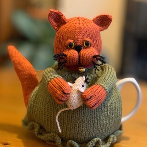 May include: A knitted orange cat teapot cosy with a green dress and a white knitted mouse in its paws.