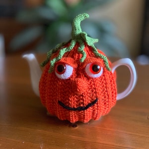 Puede incluir: Calienta-tetera de punto naranja con una cara sonriente, ojos saltones y un tallo verde en la parte superior.