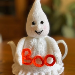 Puede incluir: Una funda de tetera de fantasma de punto blanco con una cara sonriente y la palabra "BOO" en hilo naranja.
