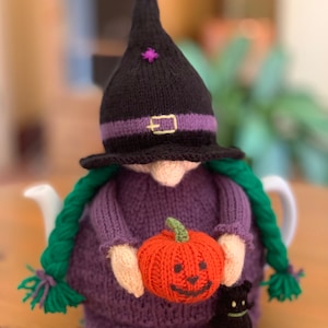 May include: A purple knitted witch tea cozy with a black hat, green hair, and a small orange pumpkin with a jack-o'-lantern face. The witch is holding the pumpkin and a black cat is sitting next to her.