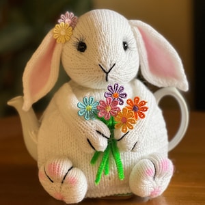 May include: White knitted bunny teapot cosy with pink ears and a bouquet of colourful embroidered flowers.