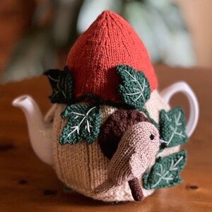 Puede incluir: Una funda de tetera de punto con un pájaro, hojas y un gorro rojo de punto. La funda es beige con detalles marrones y hojas verdes.