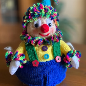 May include: A handmade knitted clown tea cozy. The cozy features a yellow shirt, blue overalls, and a colorful, textured wig and collar. It has a red nose, button eyes, and a smiling mouth. The cozy is designed to fit a teapot.