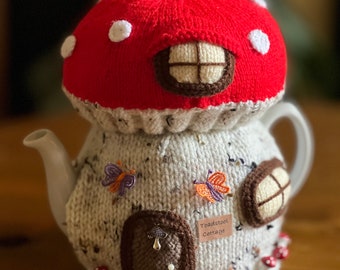 May include: A knitted teapot cosy shaped like a mushroom house with a red and white top, a white body, and brown details. The cosy has a small window, a door, and a sign that reads "Toadstool Cottage".