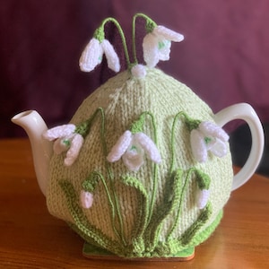 Può includere: Una coperta per teiera lavorata a maglia verde chiaro, ornata da fiori di bucaneve bianchi e verdi. La coperta è progettata per adattarsi a una teiera bianca, con i fiori disposti sulla superficie. La coperta ha un aspetto strutturato, con i fiori che aggiungono un effetto tridimensionale.