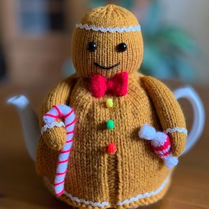 May include: A knitted gingerbread man tea cosy in shades of brown, red, and white. The cosy has a red bow tie, colourful buttons, and holds a candy cane and a wrapped sweet. The cosy is placed on a white teapot.
