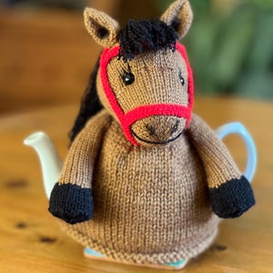 Op de afbeelding: Een bruine gebreide paard theemuts met een rode neusriem en zwarte manen en hoeven. De theemuts is ontworpen om over een witte theepot te passen, wat een grillige toets toevoegt aan de thee tijd.