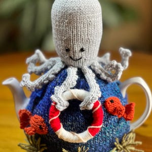 May include: A knitted tea cozy featuring a light gray octopus with black eyes sitting atop a deep blue textured base. The design includes two small orange fish, a red and white life preserver held by the octopus, and green seaweed accents.