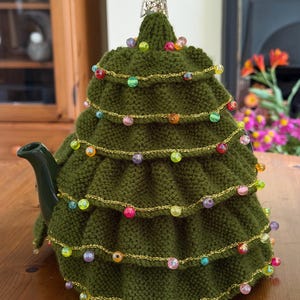 Puede incluir: Un calentador de té de árbol de Navidad tejido en verde con una estrella plateada en la parte superior. El calentador está decorado con cuentas de colores y una cuerda dorada, que se asemeja a luces navideñas. Diseñado para una tetera.
