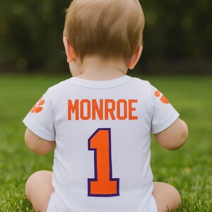 May include: A white baby bodysuit with the name "Monroe" in orange block letters and the number "1" in orange with a purple outline. The bodysuit has orange paw prints on the sleeves. The baby is sitting on green grass.