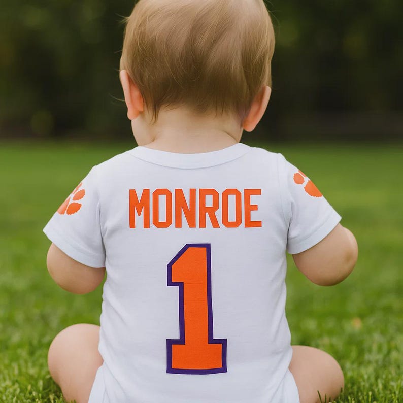 May include: A white baby bodysuit with the name "Monroe" in orange block letters and the number "1" in orange with a purple outline. The bodysuit has orange paw prints on the sleeves. The baby is sitting on green grass.
