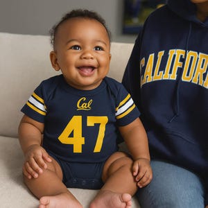 A navy blue baby bodysuit with yellow and white stripes on the sleeves. The bodysuit has the word 'Cal' above the number '47' in yellow. A person is wearing a navy blue hoodie with the word 'CALIFORNIA' in yellow.