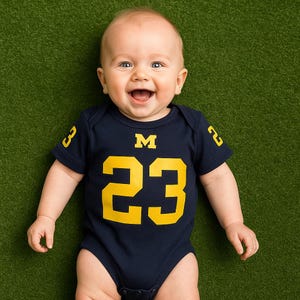 happy baby laying on grass backdrop wearing navy blue onesie with yellow 23 on body below the Michigan Wolverines logo and numbers on sleeves in yellow.