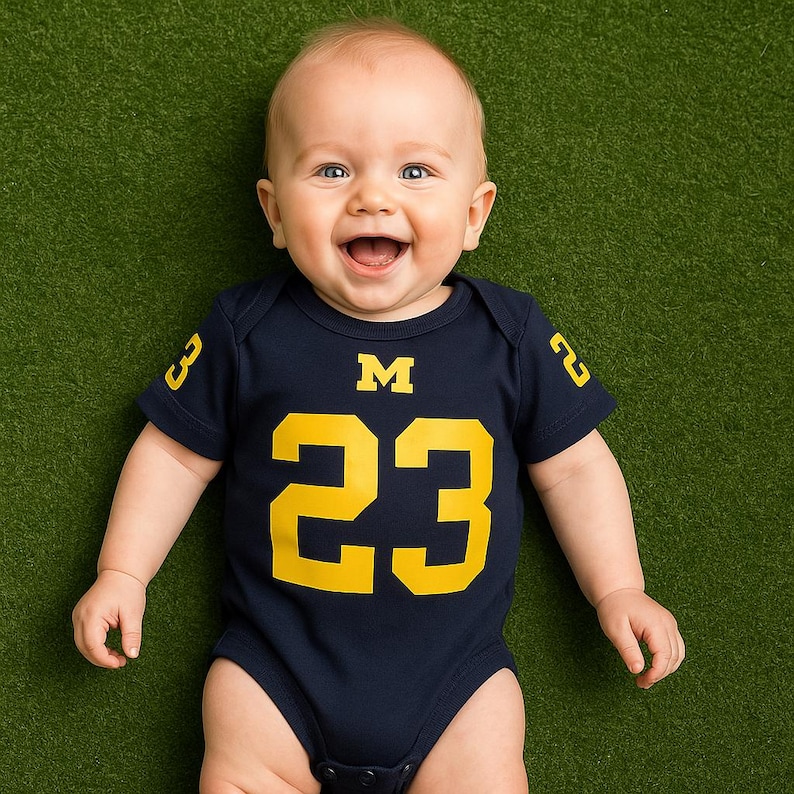 happy baby laying on grass backdrop wearing navy blue onesie with yellow 23 on body below the Michigan Wolverines logo and numbers on sleeves in yellow.